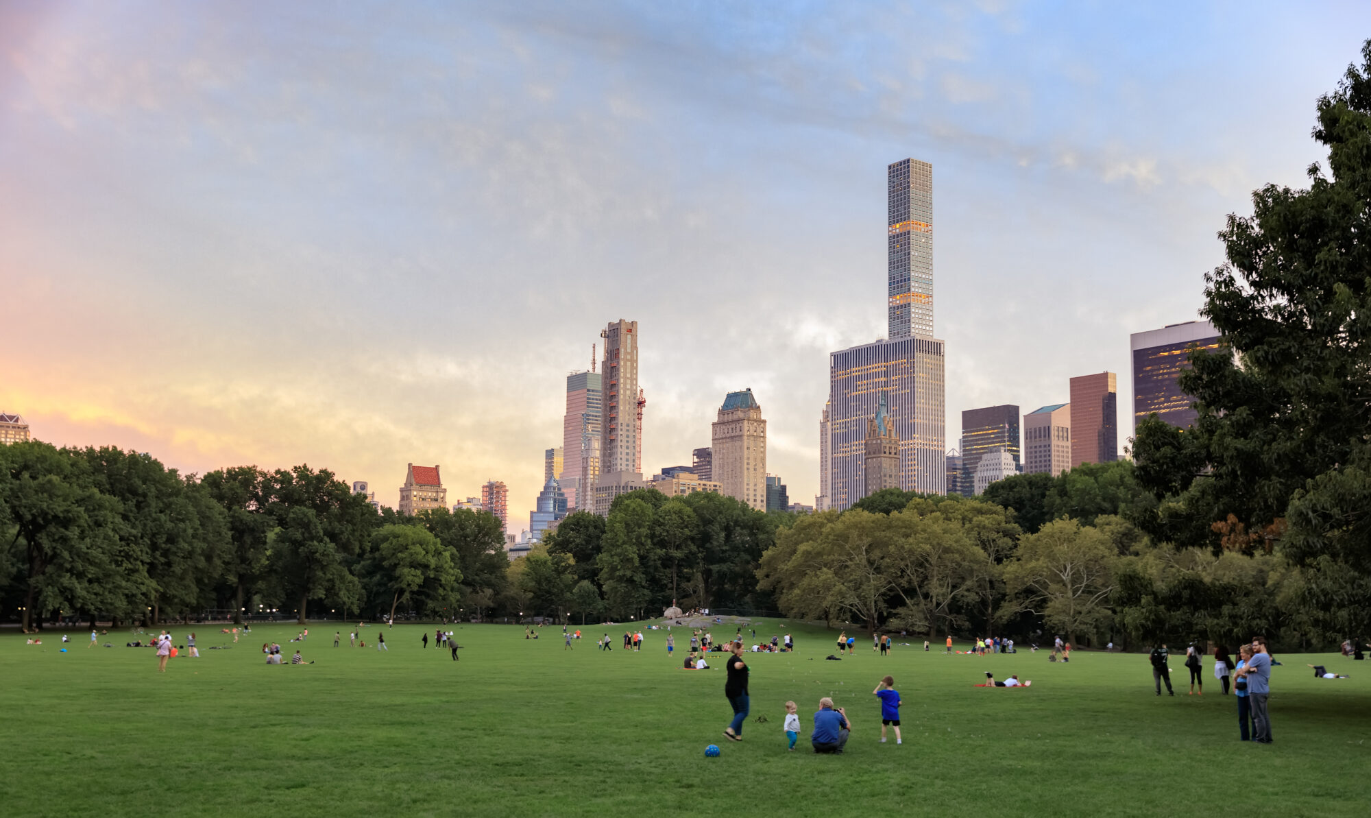 The expansive green fields of Central Park in New York City are filled with people enjoying a tranquil evening. Children play soccer, couples sit on the grass, and individuals walk or take photographs, all basking in the well-being that the park provides. In the background, the skyline features an array of skyscrapers glowing with the warm hues of the setting sun. The contrast between the natural park space and the bustling city creates a harmonious balance, emphasizing the park as a haven of well-being in the urban landscape.