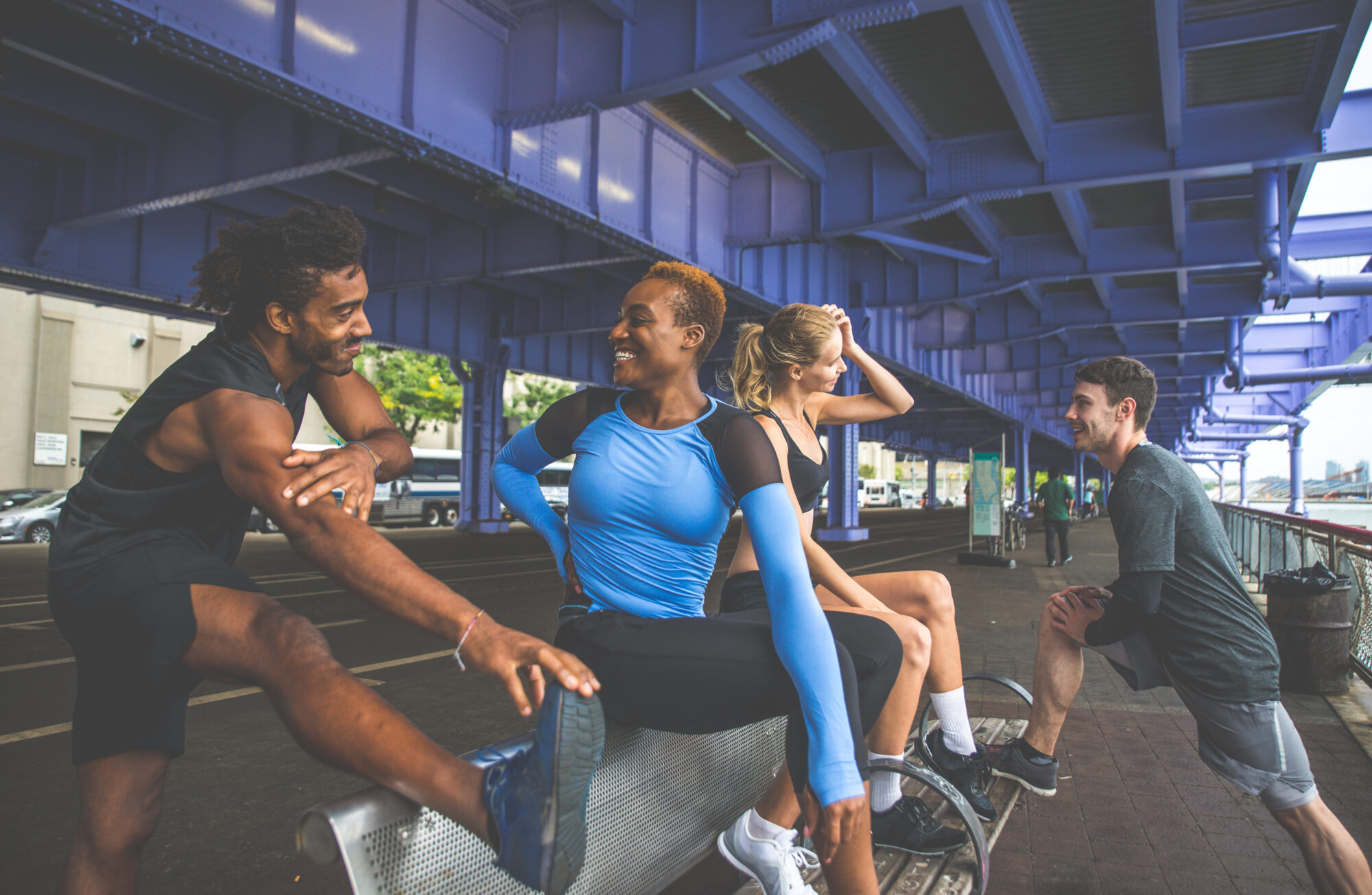 A vibrant group of runners stretching and interacting under a city bridge, embodying urban fitness and camaraderie in New York City.