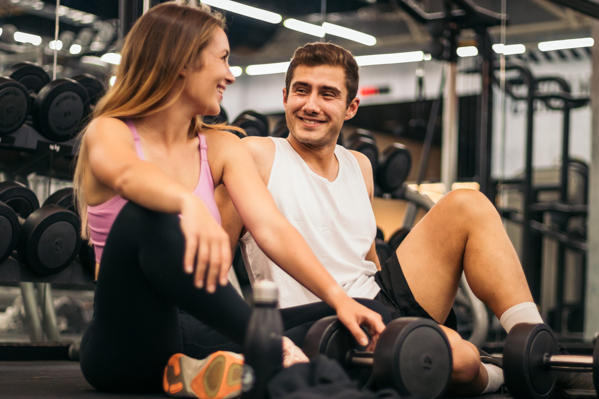 Man and woman sitting on the gym floor, smiling at each other after a workout, with weights around them, illustrating a light and natural interaction in a fitness setting.