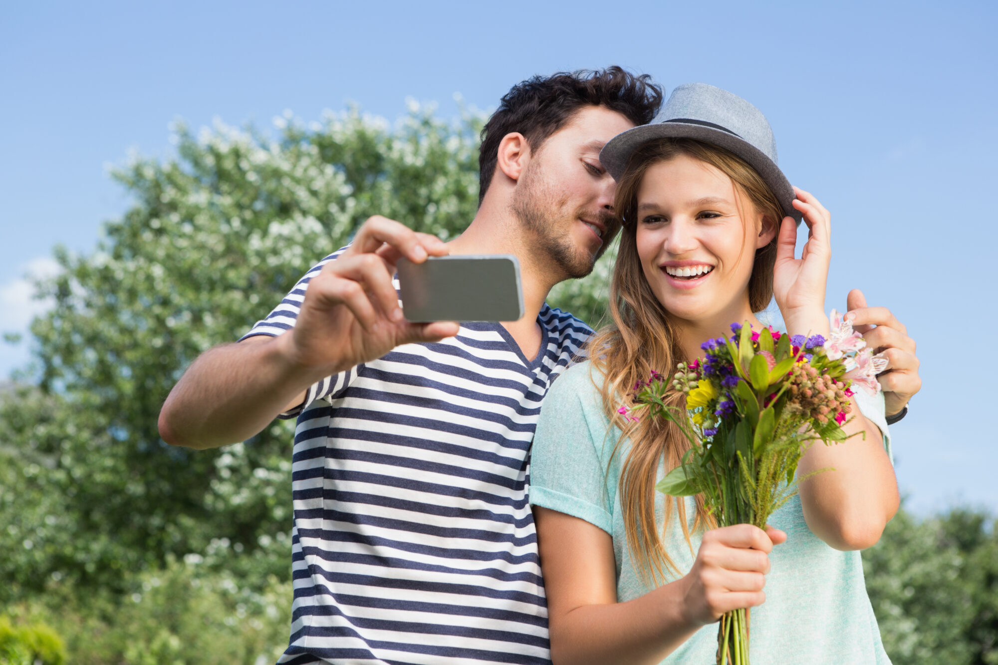 Man taking a selfie with a smiling woman holding a bouquet of flowers, as he leans in to whisper something, creating a playful and affectionate moment outdoors.